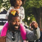 A joyful African American father with curly-haired daughter on shoulders, outdoors.